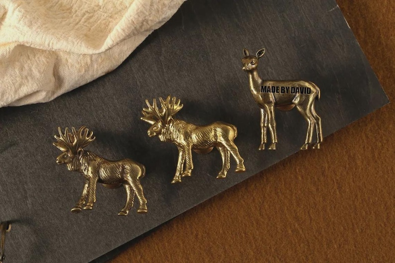 May include: Three brass animal figurines: two moose and a deer. The moose have large antlers, while the deer stands gracefully. The text "MADE BY DAVID" is on the deer figurine. The figurines are on a dark gray surface.