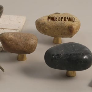 May include: A collection of stone cabinet knobs in various colors, including brown, black, and tan. Each knob is mounted on a brass base. The text "MADE BY DAVID" is visible on one of the knobs.