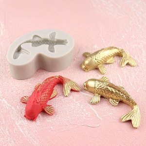 May include: A silicone mould for making cake decorations shaped like koi fish. The mould is grey and has two fish shapes. There are three finished koi fish, two gold and one red, on a pink surface.