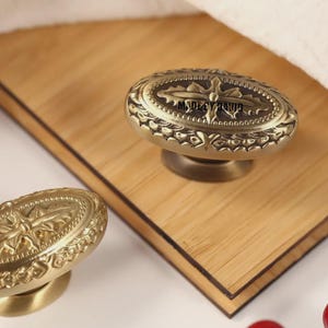 May include: Antique brass oval cabinet knobs with intricate floral and geometric detailing. The knobs are mounted on a light brown wooden surface. The text "MADE BY RONIA" is visible on the knob.