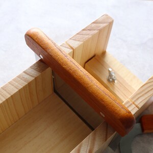 Puede incluir: Un molde de jab&oacute;n de madera con un alambre de corte de metal y un mango de madera. El molde est&aacute; hecho de madera clara y tiene una forma rectangular.