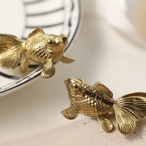 Könnte beinhalten: Zwei goldfarbene Goldfischfiguren. Die Fische haben detaillierte Schuppen, Flossen und Schwänze. Ein Fisch befindet sich auf einem weißen Teller mit schwarzen Streifen, der andere auf einer hellen Oberfläche. Der Text "MADE BY DAVID" ist auf einem der Fische zu sehen.