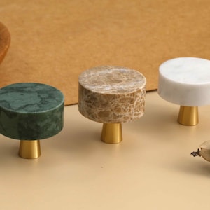 May include: Three cylindrical cabinet knobs featuring flat circular tops made of green, beige, and white marble. Each knob is attached to a small, tapered, gold-toned metal base. The knobs are arranged in a row, showcasing distinct stone patterns.