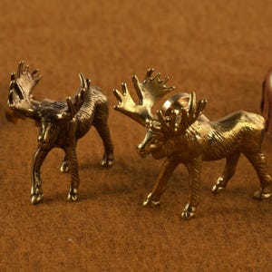 May include: Two small, detailed brass moose figurines with large antlers stand on a brown surface. One is a darker bronze color, and the other is a bright gold. A wooden slice is in the background.