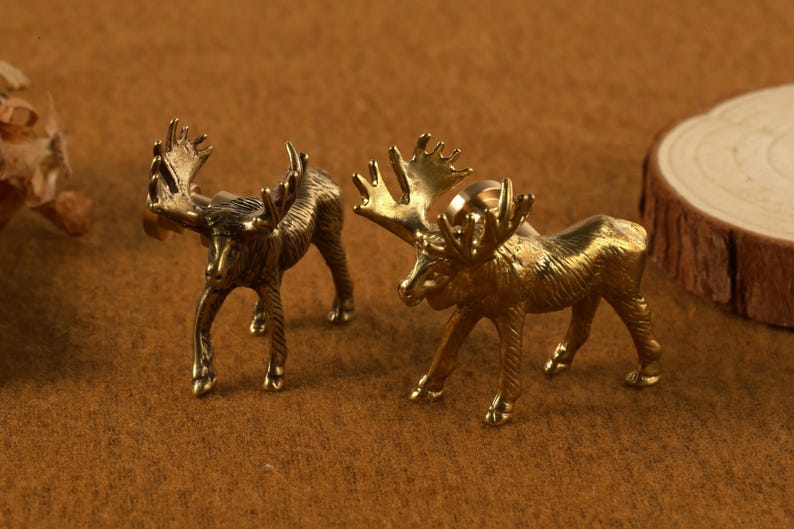 May include: Two small, detailed brass moose figurines with large antlers stand on a brown surface. One is a darker bronze color, and the other is a bright gold. A wooden slice is in the background.