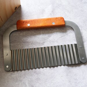 Puede incluir: Un cuchillo de acero inoxidable con borde ondulado y mango de madera. El cuchillo est&aacute; dise&ntilde;ado para cortar verduras en formas decorativas.