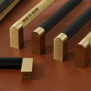 Puede incluir: Primer plano de varios tiradores de armario de latón con mangos texturizados negros y dorados. Uno de los tiradores está grabado con "MADE BY DAVID". Los tiradores están colocados sobre una superficie de cuero marrón.