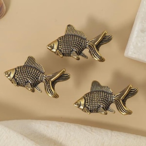 Könnte beinhalten: Drei bronzefarbene, fischförmige Dekorationsgegenstände. Jeder Fisch hat detaillierte Schuppen und Flossen mit strukturiertem Aussehen. Die Fische sind auf einer neutral getönten Oberfläche angeordnet, was auf ein nautisches oder aquatisches Thema hindeutet.