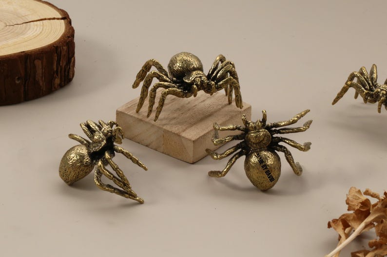 以下が含まれることがあります： 金色のクモの置物のコレクション。足や体に詳細な装飾が施されています。1匹のクモが小さな木のブロックの上に置かれています。これらのクモは、装飾品や収集品として使用される可能性があります。
