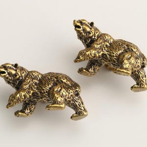 May include: Two antique brass bear figurines. The bears are depicted in a walking pose, with detailed fur texture and open mouths. The figurines have a golden-brown color and are set against a white background.