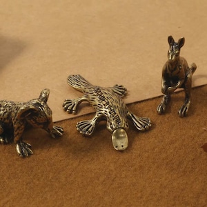 Puede incluir: Tres pequeñas figuras de animales de color bronce: un wombat, un ornitorrinco y un canguro. El wombat está agachado, el ornitorrinco está plano y el canguro está de pie. Las figuras están sobre una superficie marrón.