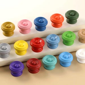 Puede incluir: Una colección de diecisiete pomos redondos de madera en varios colores sólidos, incluyendo amarillo, rosa, azul, naranja, verde oscuro, gris, rojo, azul claro, verde brillante, madera natural, morado, granate, cerceta y blanco, con un detalle circular.