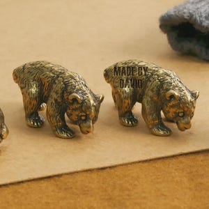 Può includere: Tre piccole statuette di orso color bronzo. Gli orsi sono in posizione di camminata, con una texture di pelliccia dettagliata. Il testo "MADE BY DAVID" è visibile su un orso. Le statuette sono su una superficie marrone.