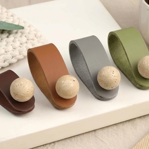 May include: Four leather drawer pulls in dark brown, tan, grey, and olive green. Each pull is a folded loop of leather secured by a round, speckled, beige stone knob. The pulls are arranged in a row, showcasing their distinct colours and textures.