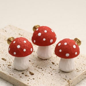 May include: Three mushroom-shaped cabinet knobs featuring red caps with white polka dots and white stems. Each knob has a textured, hand-carved appearance and includes a brass-colored metal mounting screw attached to the back of the cap.