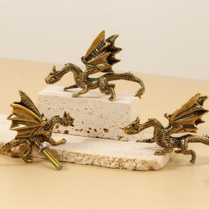 Può includere: Tre figurine di drago color oro antico con ali spiegate sono esposte su una superficie colorata chiara. Un drago ha un'asta color oro attaccata. I draghi sono su una superficie chiara e strutturata. Il testo "MADE BY DAVID" è visibile sull'ala di un drago.