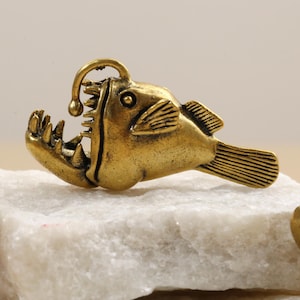 May include: A brass-coloured anglerfish figurine with an open mouth, sharp teeth, and a glowing lure. The fish is resting on a white, textured surface. The figurine has detailed fins and a textured body.