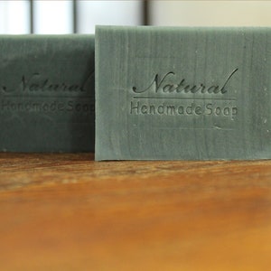 Op de afbeelding: Twee rechthoekige handgemaakte zeepblokken in een gedempte groene tint. De zeepblokken zijn voorzien van de tekst "Natural Handmade Soap". De afbeelding is een close-up, die de textuur en details van de zeep benadrukt.
