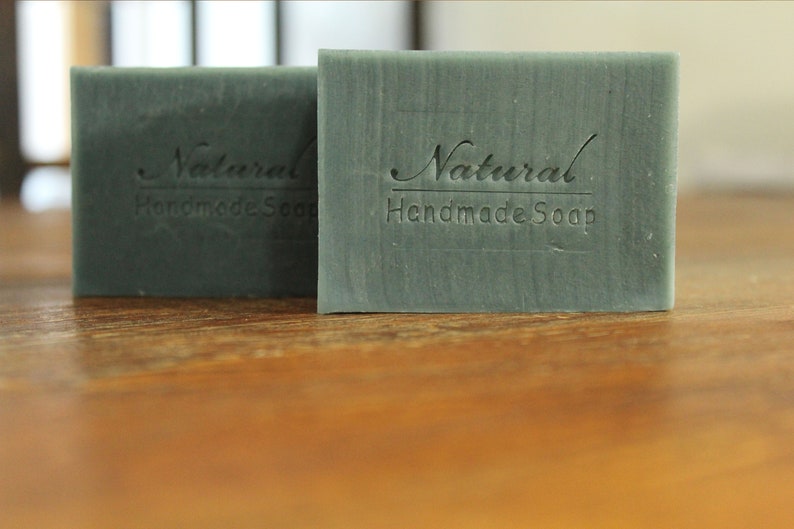 Op de afbeelding: Twee rechthoekige handgemaakte zeepblokken in een gedempte groene tint. De zeepblokken zijn voorzien van de tekst "Natural Handmade Soap". De afbeelding is een close-up, die de textuur en details van de zeep benadrukt.