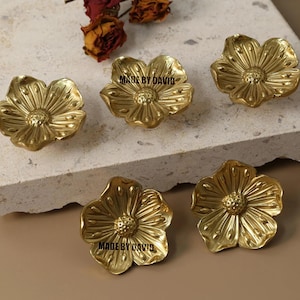 Könnte beinhalten: Sechs goldfarbene Schrankknöpfe in Blumenform. Jeder Knopf hat ein detailliertes Blütenblattdesign und einen strukturierten Kern. Der Text "MADE BY DAVID" ist auf zwei der Knöpfe zu sehen. Die Knöpfe sind auf einer hellen Oberfläche angeordnet.