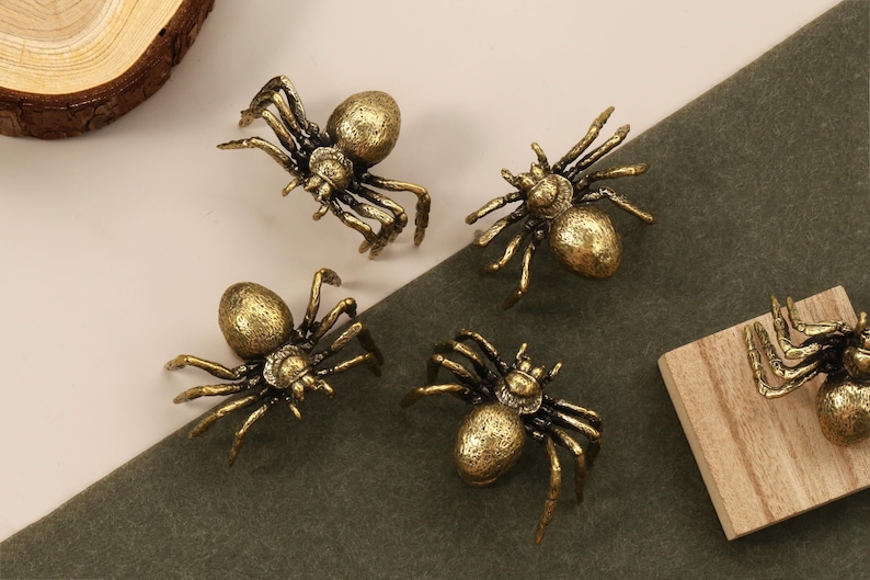 May include: Close-up of several antique gold-colored spider-shaped cabinet knobs. The knobs have detailed legs and a textured body. The spiders are arranged on a neutral background, suggesting a decorative hardware theme.