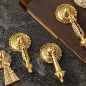 May include: Close-up of several antique brass drawer pulls. The pulls have a circular backplate with ornate detailing and a decorative handle. The text "MADE BY DAVID" is engraved on the handle of some of the pulls. The pulls are a warm gold colour.
