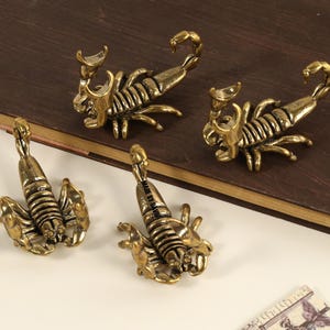 May include: Four antique brass scorpion figurines. The scorpions are detailed with segmented bodies, claws, and curved tails. They are arranged on a dark brown surface with a light-coloured surface in the foreground.