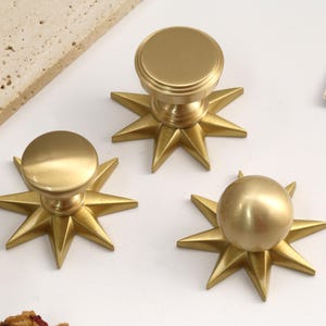 May include: Three gold-toned metal cabinet knobs, each featuring a circular top and a starburst-shaped base with eight pointed rays. The knobs have a smooth, matte finish and are arranged in a cluster on a white surface.