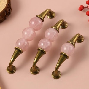 May include: Three cabinet pulls featuring antique brass-toned metal hardware and spherical light pink stone accents. The pulls vary in length, with the largest having three stone spheres, the middle having two, and the smallest having one sphere.