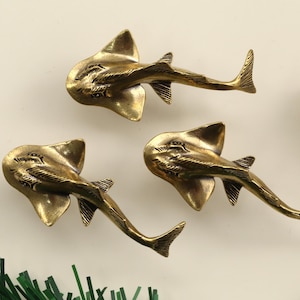 May include: Three brass-colored, stylized ray fish figurines with detailed fins and tails. The figurines are arranged on a light background, near a seashell, a piece of white stone, and some green decorative elements. The rays are approximately 4 inches long.