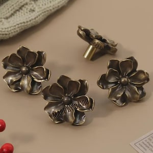 May include: Four bronze-colored flower-shaped cabinet knobs. One knob is attached to a brass stem. The knobs are arranged on a neutral surface with a sprig of red berries and a knitted textile in the background. The text "MADE BY DAVID" is visible on one knob.