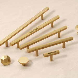 Puede incluir: Un conjunto de herrajes dorados para armarios, que incluyen tiradores y pomos. Los tiradores tienen un diseño hexagonal y el texto "MADE BY DAVID" está impreso en algunos. Los pomos tienen forma octogonal. Medidas en centímetros.