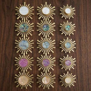 May include: A collection of fifteen sunburst-shaped cabinet knobs arranged in three columns. Each knob features a central circular inlay made of various iridescent materials, including white, abalone, blue, purple, and peach tones, set in gold metal.