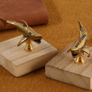 Könnte beinhalten: Zwei messingfarbene, haiförmige Objekte, jeweils auf einem kleinen, quadratischen Holzsockel montiert. Ein Hai trägt den Text "MADE BY DAVID" auf der Seite. Die Haie sind mit Flossen, Kiemen und offenem Maul detailliert.