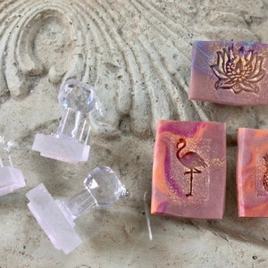 Op de afbeelding: Ambachtelijke zepen met ingebedde ontwerpen, waaronder een lotusbloem, een flamingo en een ananas. De zepen hebben een roze, oranje en paars gemarmerd effect. Heldere, facetgeslepen stempels liggen verspreid rond de zepen.