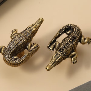 May include: Two antique brass-colored alligator-shaped cabinet pulls. The detailed reptile figures are positioned on a light beige surface. The pulls are designed with intricate scales and small feet, adding a unique touch to furniture.