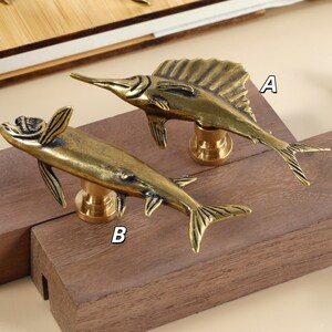 Op de afbeelding: Twee messing kastknoppen in de vorm van een vis, een met een zeilachtige vin en de andere met kleinere vinnen. De knoppen zijn gemonteerd op een houten blok, wat hun ontwerp laat zien.