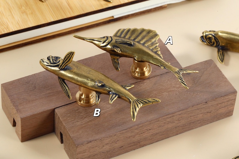 Op de afbeelding: Twee messing kastknoppen in de vorm van een vis, een met een zeilachtige vin en de andere met kleinere vinnen. De knoppen zijn gemonteerd op een houten blok, wat hun ontwerp laat zien.