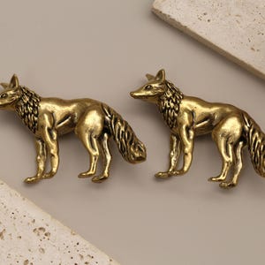 Op de afbeelding: Twee messing vossenbeeldjes in een staande pose, met gedetailleerde vachttextuur. De vossen zijn goudkleurig en staan op een neutrale achtergrond met decoratieve stenen accenten. Een derde vossenbeeldje is zichtbaar op de achtergrond.