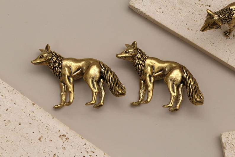 Op de afbeelding: Twee messing vossenbeeldjes in een staande pose, met gedetailleerde vachttextuur. De vossen zijn goudkleurig en staan op een neutrale achtergrond met decoratieve stenen accenten. Een derde vossenbeeldje is zichtbaar op de achtergrond.