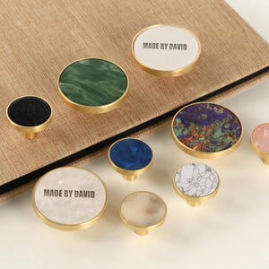 Può includere: Una collezione di pomelli decorativi per mobili con vari disegni e colori, tra cui motivi in marmo verde, blu e bianco. Alcuni pomelli hanno la scritta "MADE BY DAVID". Ogni pomello ha una base dorata.