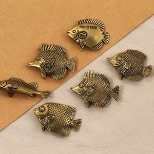 May include: A collection of bronze-coloured fish-shaped decorative objects. One fish has the text "MADE BY DAVID" on it. The fish vary in size and design, with detailed scales and fins. They are arranged on a two-tone background.