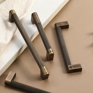May include: Bronze cabinet pulls with textured handles and rectangular ends. Some pulls feature a cat silhouette and the names "Luna" and "Milo". The pulls are arranged on a neutral surface with a white fabric and green leaf accents.