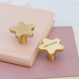 May include: Two star-shaped brass cabinet knobs rest on a pink and purple notebook. One knob has the text "MADE BY DAVID". The knobs have a gold finish and are mounted on a cylindrical base.
