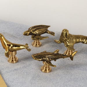 Könnte beinhalten: Vier Tierfiguren aus Messing: eine Robbe, eine Schildkröte, ein Walross und ein Hai. Jede Figur ist auf einem kurzen, runden Sockel montiert. Die Robbe trägt den Text "MADE BY DAVID" auf ihrer Seite. Die Figuren sind goldfarben und detailliert.