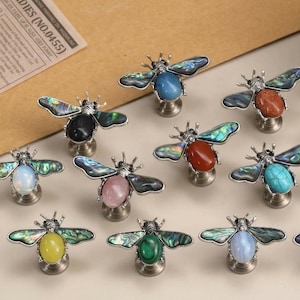 Può includere: Una collezione di pomelli per mobili a forma di ape con ali in conchiglia di abalone iridescente e cabochon ovali centrali in vari colori, tra cui blu, arancione, bianco, rosa, rosso, turchese, giallo, verde e blu scuro, su basi metalliche.
