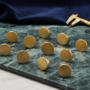 Puede incluir: Diez pomos de armario redondos de latón martillado, dispuestos sobre una superficie de mármol verde y gris. Un tejido azul oscuro y un calibrador de latón están en el fondo. Los pomos tienen una superficie texturizada.