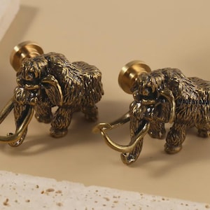 May include: Two bronze-coloured cabinet knobs shaped like woolly mammoths. The knobs have a detailed, textured finish and curved tusks. The text "MADE BY DAVID" is visible on the right knob. These decorative knobs are suitable for furniture.