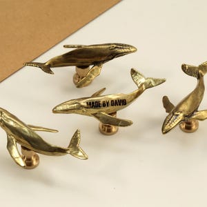 May include: Four brass whale-shaped cabinet knobs. Each whale is detailed with fins and a tail, mounted on a small, round base. The text "MADE BY DAVID" is on one whale. The knobs are on a white surface.
