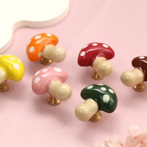Pode incluir: Um conjunto de seis puxadores de armário em forma de cogumelo com topos brilhantes em amarelo, laranja, rosa, vermelho, verde escuro e castanho. Cada topo tem pintas brancas e um caule bege claro fixado numa base metálica cor de latão.